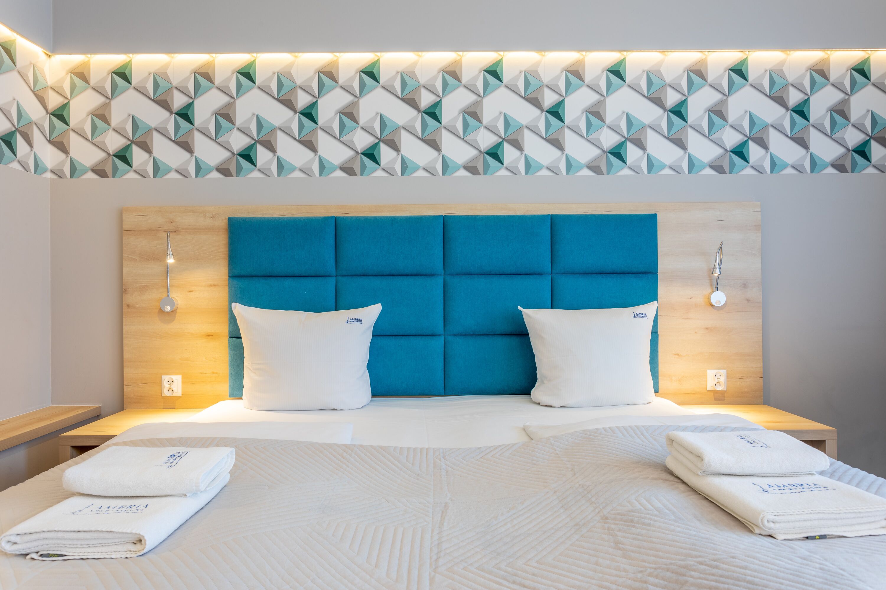 Ein Hotelzimmer mit modernem Design, ausgestattet mit einem blauen, gepolsterten Kopfteil, weißer Bettwäsche und einer Wand mit geometrischem Muster.