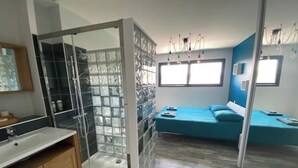 A modern bedroom with a glass shower enclosure, a bed with blue bedding, and a wooden vanity.