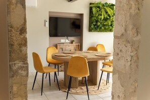 A dining area with a round wooden table, six yellow chairs, a flat-screen TV mounted on the wall, and a green leafy wall decoration. - Gîte Guyane (Eymet)