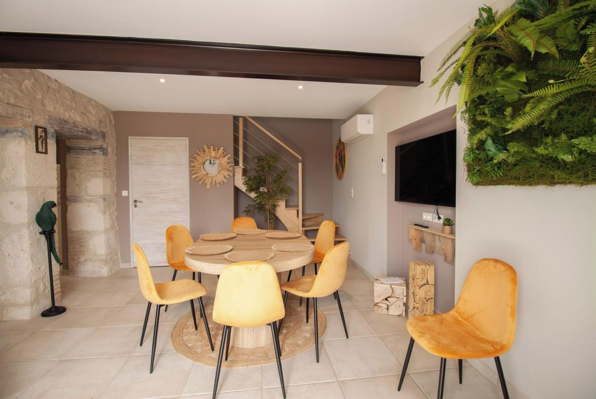 A dining area with a round table and six chairs, a staircase, a mounted TV, and a wall-mounted plant.