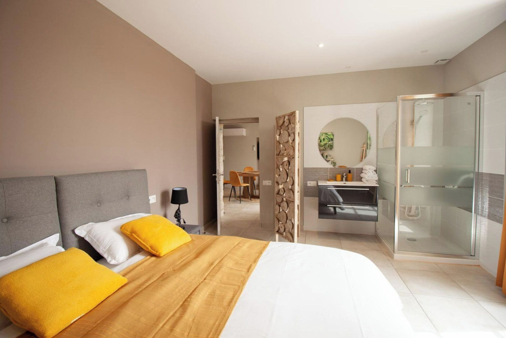 Una habitación de hotel moderna con una cama grande, un baño con ducha de vidrio y un comedor visible a través de una puerta abierta.
