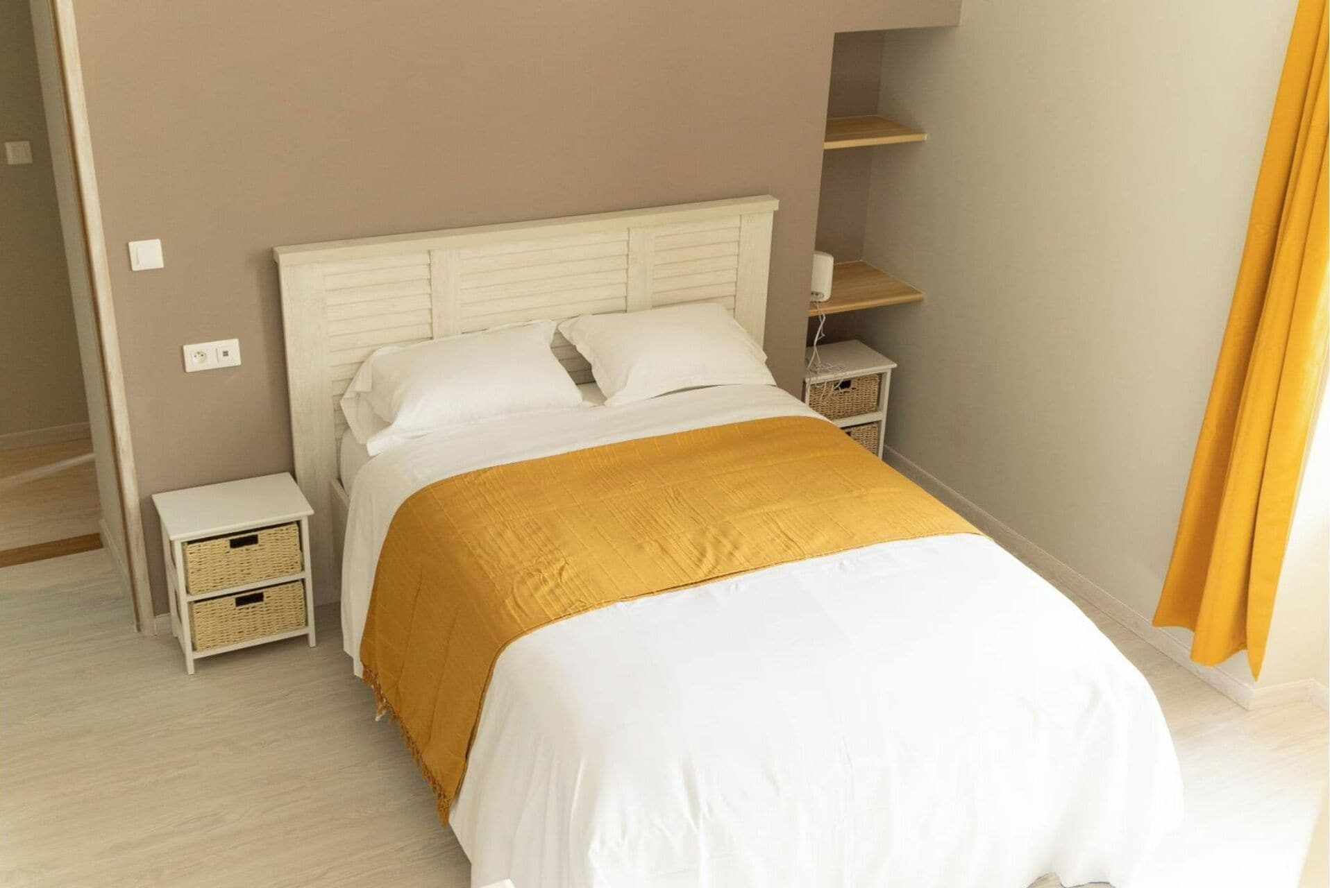 A bedroom with a white bed, a mustard-colored throw blanket, and two bedside tables with wicker baskets.