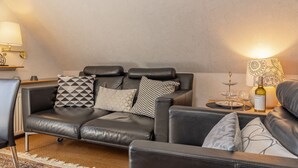 Ett modernt vardagsrum med en svart lädersoffa, mönstrade kuddar, ett litet bord med en vinflaska och en dekorativ lampa.