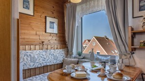 A cozy room with a wooden wall, a bench with patterned cushions, a round wooden table set with breakfast items, and a view of buildings through a large window. - The simply furnished apartment Am Seestern Carolinensiel awaits you (Carolinensiel)