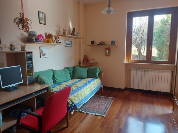 Una habitación acogedora con un escritorio de madera, una silla roja, un sofá verde y un ventanal con vista a árboles.