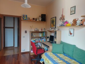 Una habitación con cama, escritorio, silla, estanterías llenas de juguetes y libros, y una puerta.