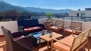 A rooftop terrace with outdoor furniture, a glass table, and a view of mountains.