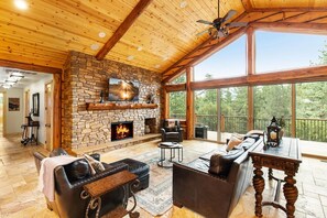 Un salón amplio con chimenea de piedra, muebles de cuero y grandes ventanas que ofrecen vistas al exterior.
