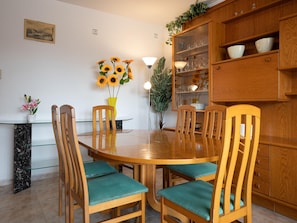 Un coin repas comprenant une table ronde en bois, six chaises en bois avec des coussins turquoise, une table basse avec un plateau en verre, un buffet en bois avec des portes en verre et un pot de fleurs contenant des tournesols.