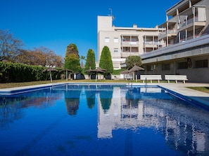 Une piscine à l’eau d’un bleu limpide, entourée de bancs et de parasols, avec un immeuble à plusieurs étages en arrière-plan.