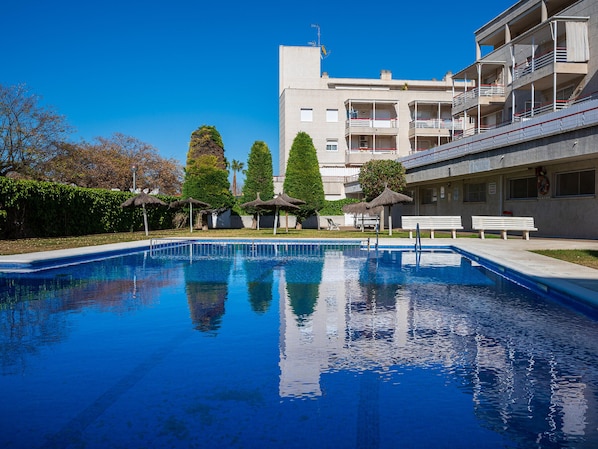 A swimming pool with clear blue water, surrounded by benches and umbrellas, with a multi-story building in the background.