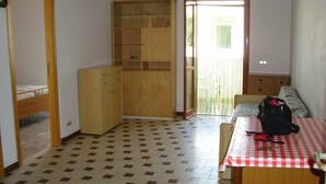 Une pièce avec un sol à carreaux, un buffet en bois, une table recouverte d’une nappe rouge et blanche, et une porte donnant sur un espace extérieur.