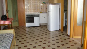 Une cuisine compacte équipée d’une cuisinière, d’un four et d’un réfrigérateur. On y trouve une petite table avec une chaise, et un lit est visible au premier plan.