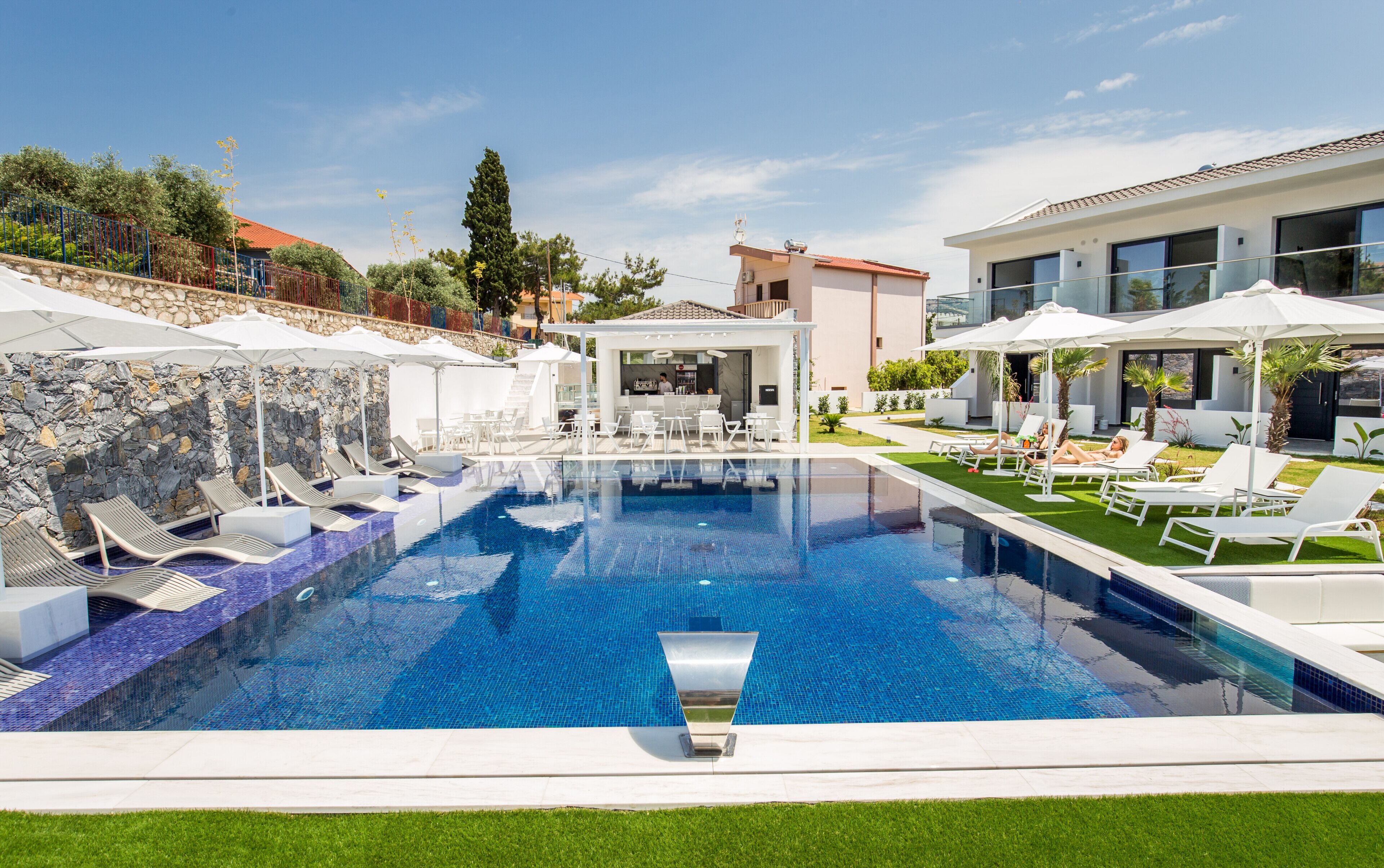 Una piscina moderna con sillas de descanso y sombrillas, rodeada de un muro de piedra y vegetación.