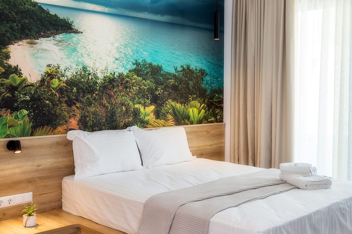 Quarto de hotel moderno com uma cama grande, vista para uma praia tropical e cabeceira de madeira.