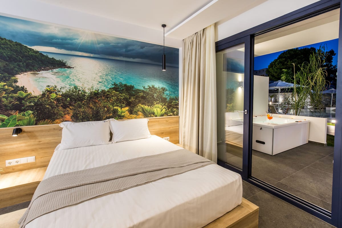 Quarto moderno com uma cama grande, vista para o mar através de uma porta de vidro e uma pequena varanda com mesa e cadeiras.
