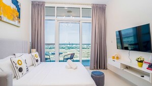 A modern hotel room with a large bed, a flat-screen TV, a balcony with a city view, and a large painting on the wall.