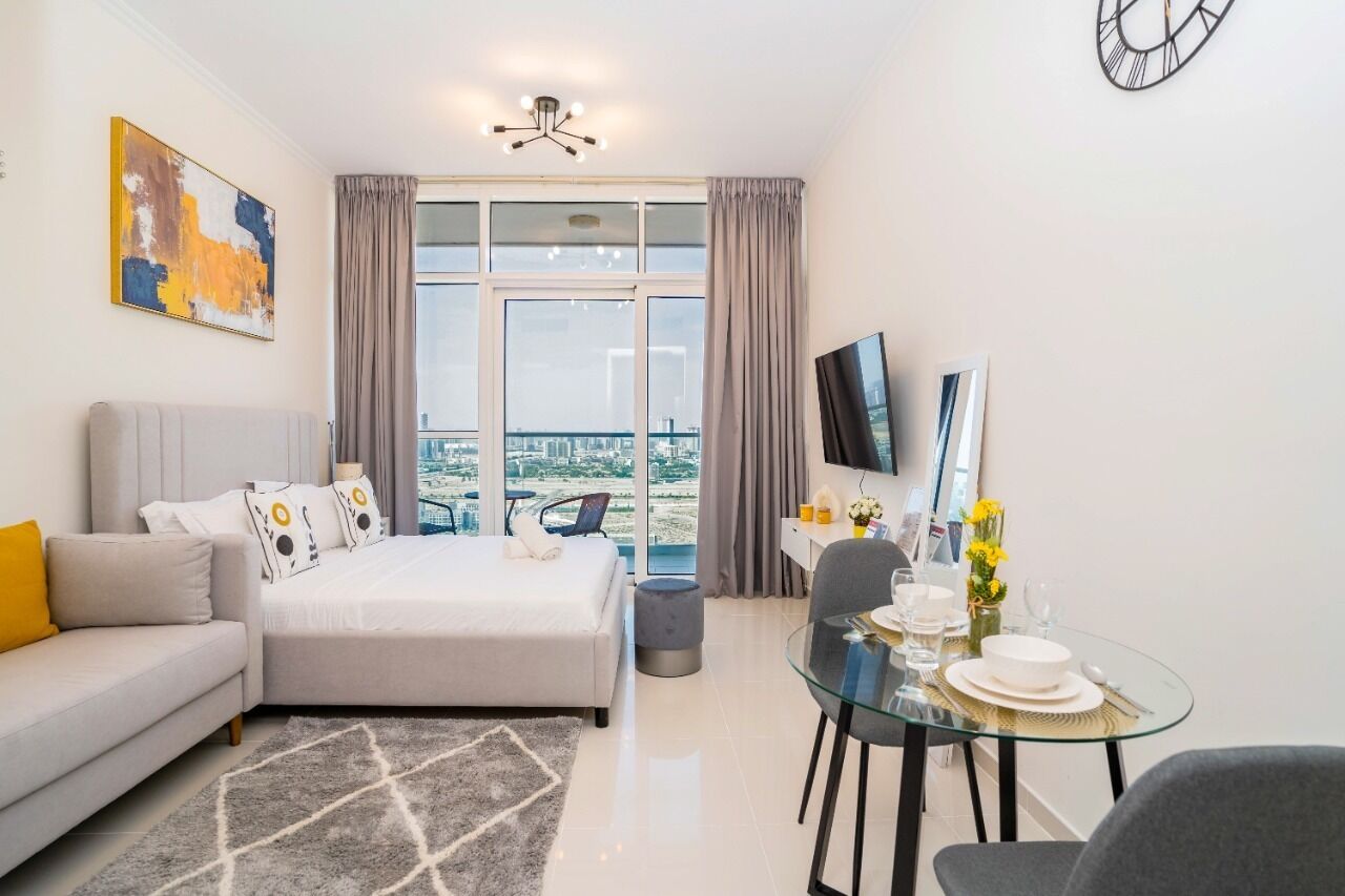 A modern hotel room with a bed, a sofa, a dining table with chairs, a TV, and a large window offering a city view.