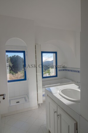 A bathroom with a bathtub, a window with a view of trees, and a tiled floor. - VILLA L'ORANGER (La Croix-Valmer)