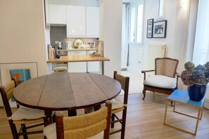A dining area with a round wooden table, four upholstered chairs, a small side table with a vase, and a kitchen in the background. - DAGUERRE MARKET, LIVELY, 30–60 days ahead? Ask for a private offer DAG-VBp (Paris)