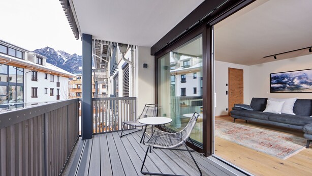 Un balcon doté d’un plancher en bois, une porte vitrée donnant sur un salon avec un canapé et un tableau, et une vue sur des bâtiments et des montagnes en arrière-plan.