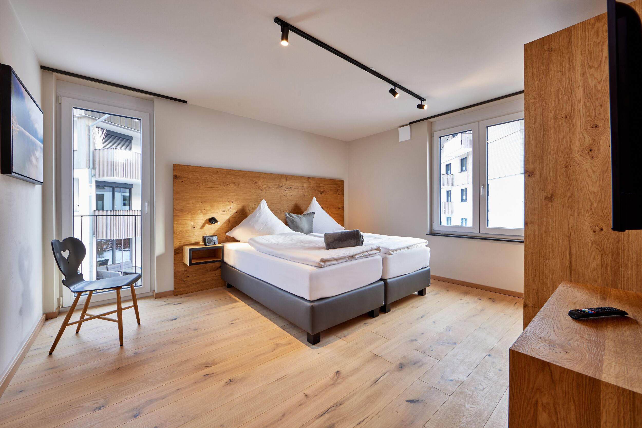 Een moderne slaapkamer met een houten hoofdbord, een bed met witte beddengoed, een nachtkastje, een stoel en een houten bureau met een afstandsbediening.