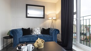 A modern living room with a blue sofa, a coffee table with mugs and a decorative item, a floor lamp, and a large window with a view of the outside. - 8 Avery Gardens (Darlington)