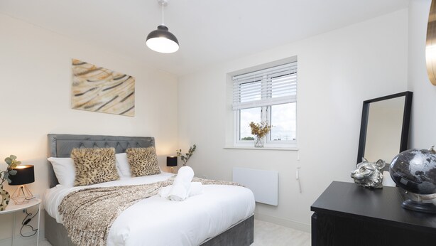 A modern bedroom with a large bed, a bedside table with a lamp, a window with blinds, and a dresser with decorative items. - 8 Avery Gardens (Darlington)