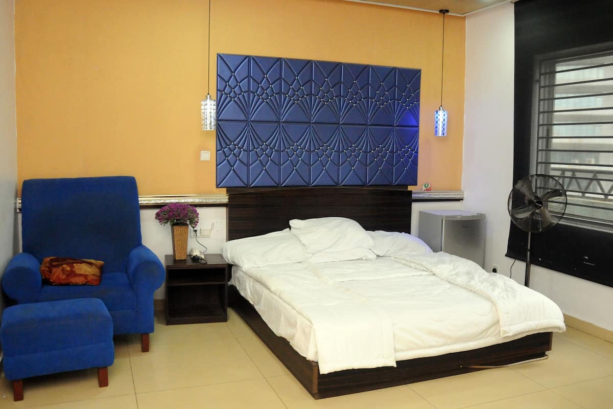 Un dormitorio con una cama, un sillón azul, una mesita de noche con un jarrón y una lámpara de pared.