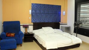 A bedroom with a bed, a blue armchair, a nightstand, a decorative wall panel, a fan, and a window with blinds. - Skywood suite  (Abeokuta)