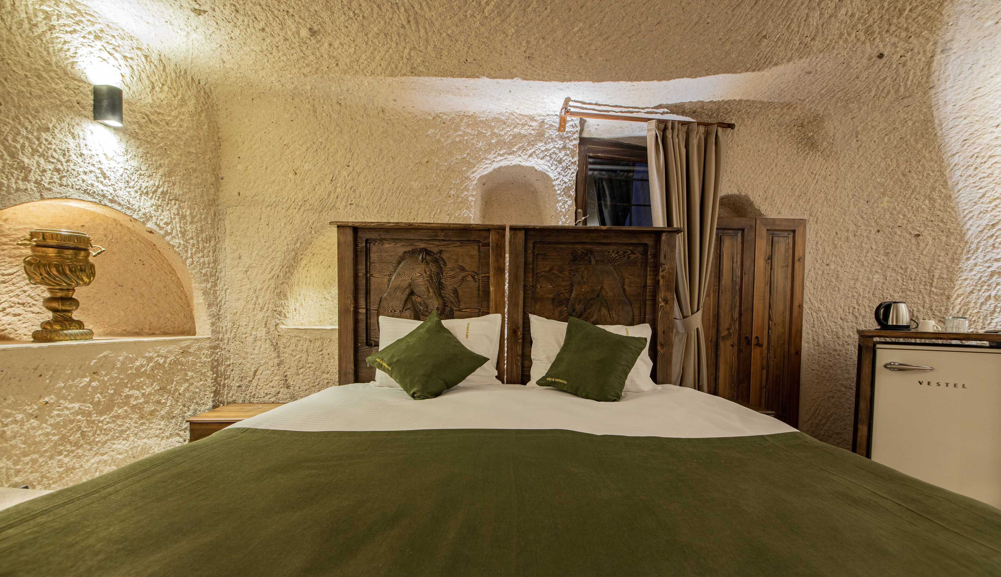 A bedroom with a rustic stone wall, a wooden bed with carved headboard, and a small bedside table with a lamp.