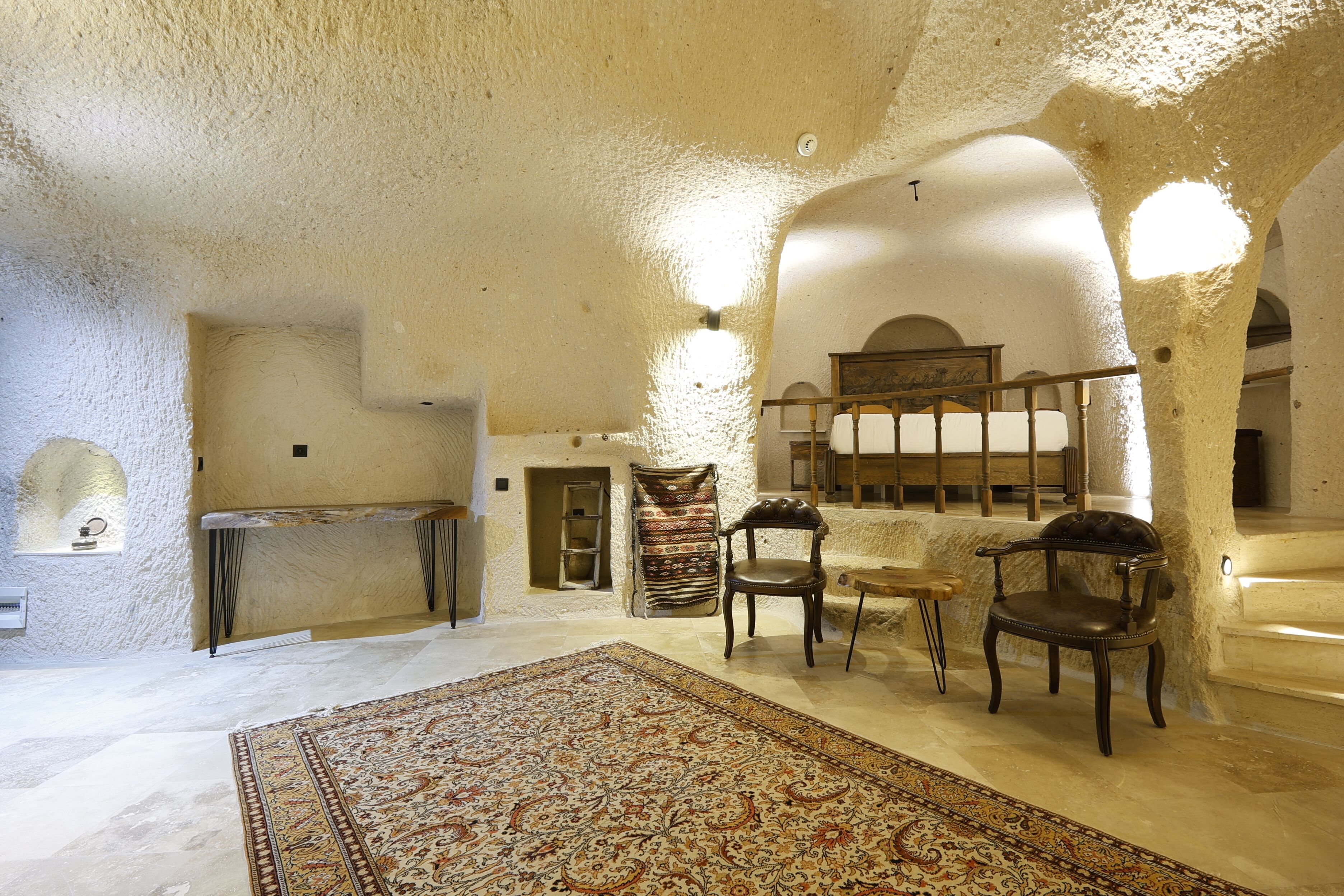 A room with a large rug, wooden chairs, a bench, and a stone wall.