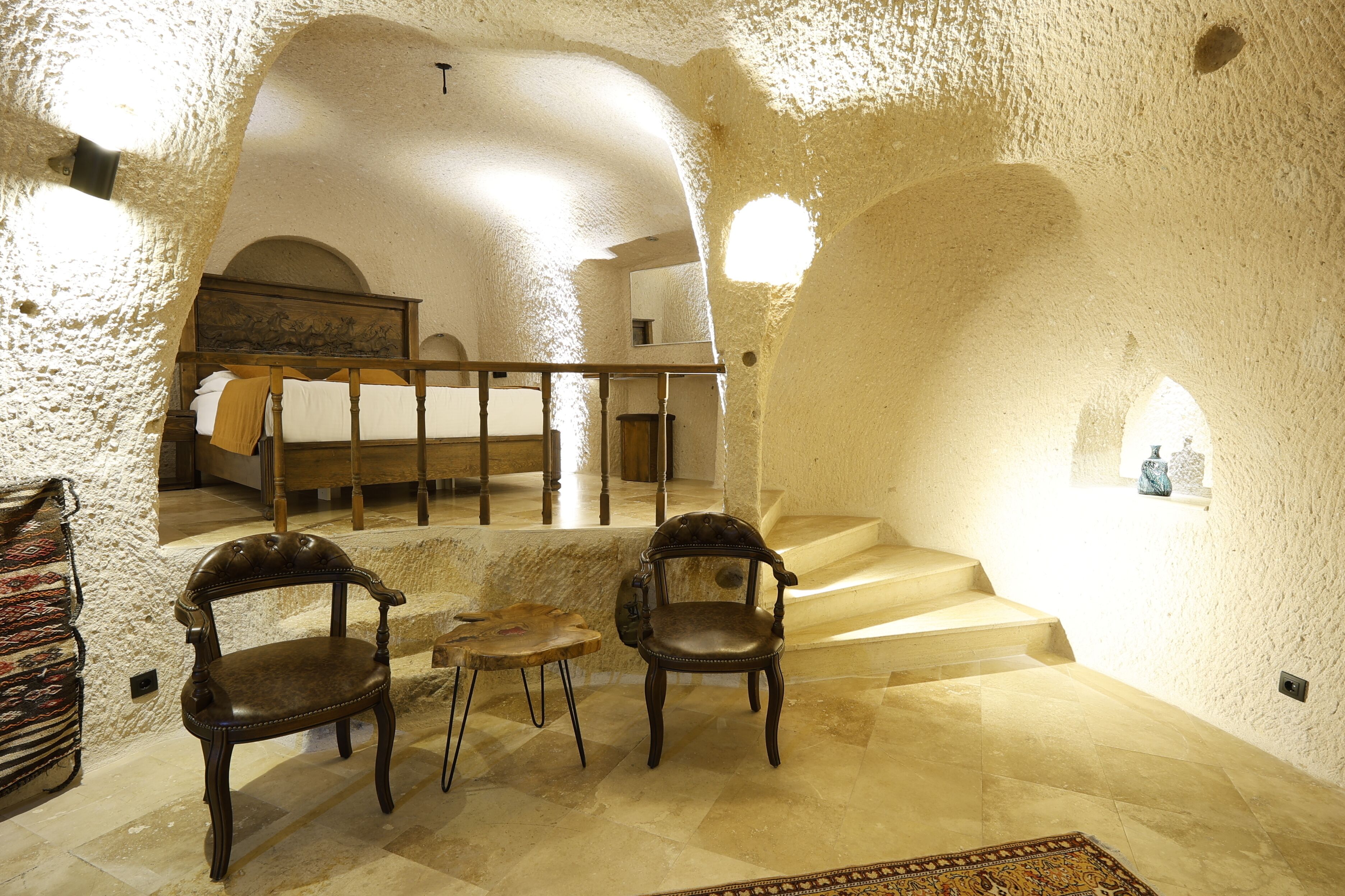 Une pièce dotée d’un plafond haut et de niches en arc de cercle, comprenant un lit, deux chaises, une petite table et un escalier.
