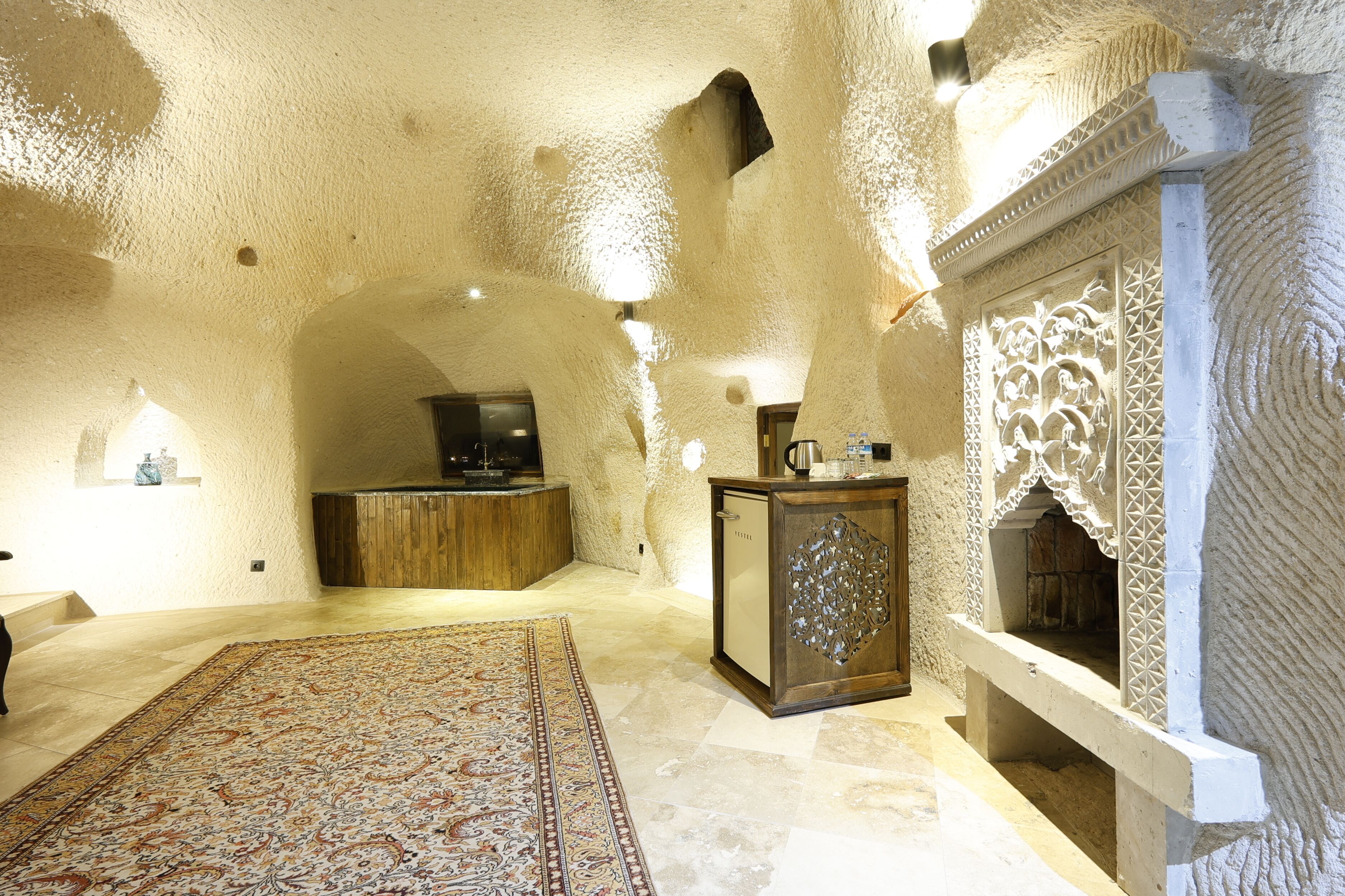 Una habitación con techo alto y texturizado, alfombra estampada, mueble de madera y chimenea decorativa.