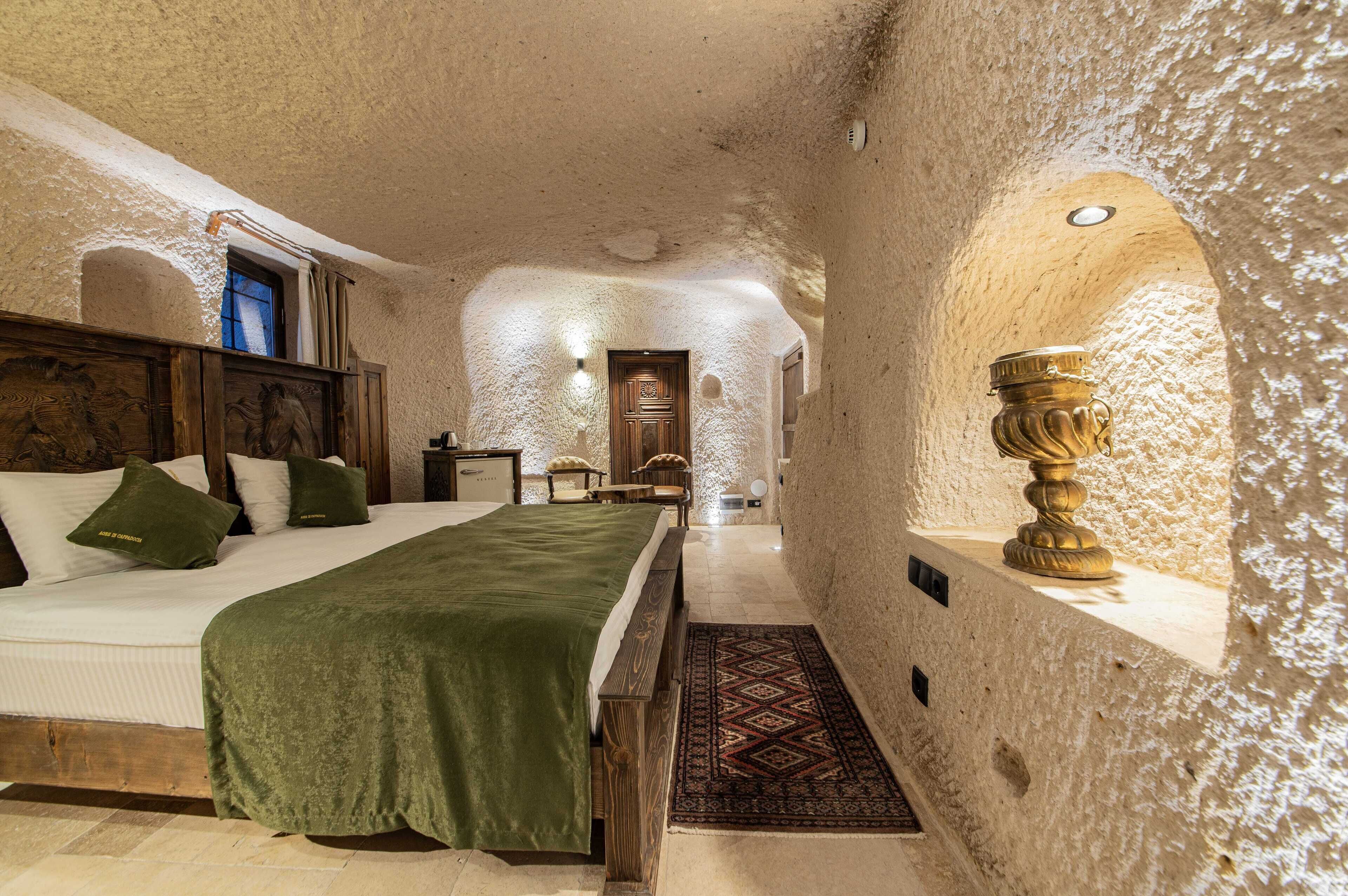 Una habitación con una cama, cabecera de madera, manta verde, alfombra estampada y un gran jarrón decorativo.