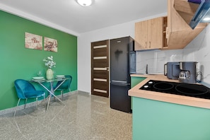 A modern kitchen with a green wall, wooden cabinets, a black refrigerator, a coffee maker, and a dining area with blue chairs and a glass table. - Akira One Bedroom Apartment (Bucharest)