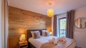 Een slaapkamer met een houten hoofdbord, een groot bed met wit beddengoed, een bedlamp, een klein houten nachtkastje, een witte vloerkleed en een raam met gordijnen.