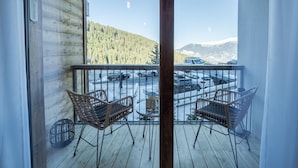 Een balkon met twee rieten stoelen, een klein tafeltje en uitzicht op een parkeerplaats met auto's en een bebost gebergte op de achtergrond.