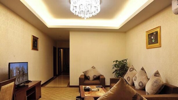 A living room with a television, sofa, and coffee table. There is a chandelier on the ceiling and a plant on the right side.
