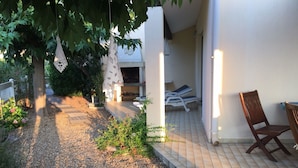 Un patio con una terraza de madera, una zona de estar con una silla de madera y una tumbona bajo un árbol.