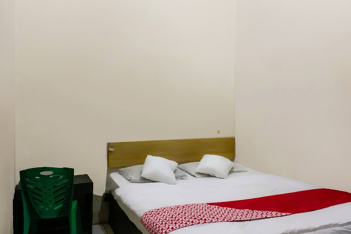 Een kleine, eenvoudige kamer met een bed, een groene plastic stoel en een houten hoofdeinde.