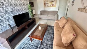 Un salón con televisor de pantalla plana, mueble de madera oscura para equipo multimedia, alfombra de cuadros, sofá color beige y pared con diseño de piedra.