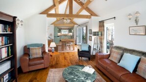 A spacious living room with a high ceiling, wooden beams, a fireplace, a dining area, and a kitchen in the background. - Long Roods Cottage, Monsal Head, near Bakewell (Bakewell)