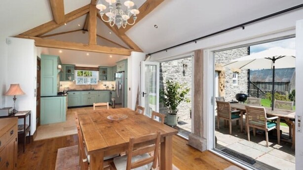 A spacious kitchen with wooden beams, a dining area with a wooden table and chairs, and a view of the garden through large sliding doors. - Long Roods Cottage, Monsal Head, near Bakewell (Bakewell)