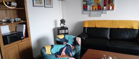 A living room with a black leather sofa, a wooden cabinet, a colorful armchair, a coffee table with decorative items, and framed artwork on the wall.
