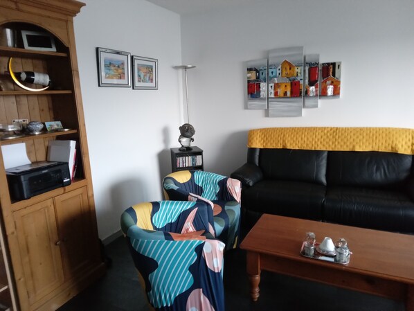 A living room with a black leather sofa, a wooden cabinet, a colorful armchair, a coffee table with decorative items, and framed artwork on the wall. - Cosy apartment in Vannes for 4 people. (Vannes)