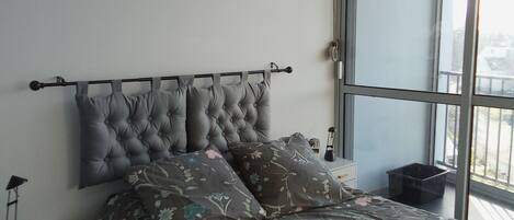 A bedroom with a tufted headboard, a floral bedspread, a bedside table with a lamp, and a large window offering a view of buildings.