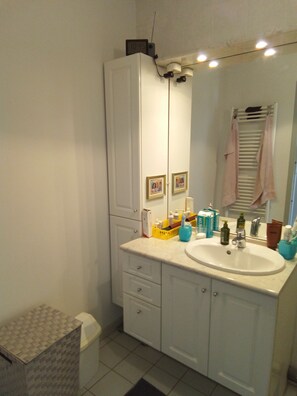 A bathroom with a white sink, mirror, and cabinets. There are toiletries on the counter and a towel rack above the mirror. - Cosy apartment in Vannes for 4 people. (Vannes)