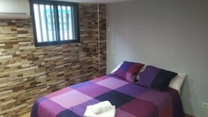 Un dormitorio con pared de piedra, una cama con colcha púrpura y azul, y una ventana con persianas.