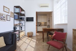 A compact living space with a sofa, a red chair, a bookshelf, a flat-screen TV, and a kitchen area visible through an open doorway. - GuestHost - Red Bridges Sunny Apartament (Napoli)
